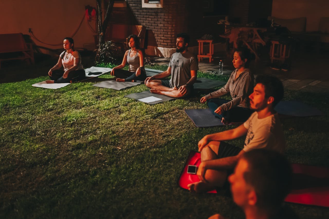 A diverse group meditating outdoors on yoga mats during evening time.